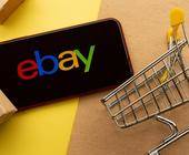 Ebay Logo auf Smartphone Screen, daneben Pakete und Einkaufswagen