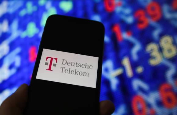 Telekom-Logo auf einem Smartphone vor einer Tafel mit Aktienkursen 